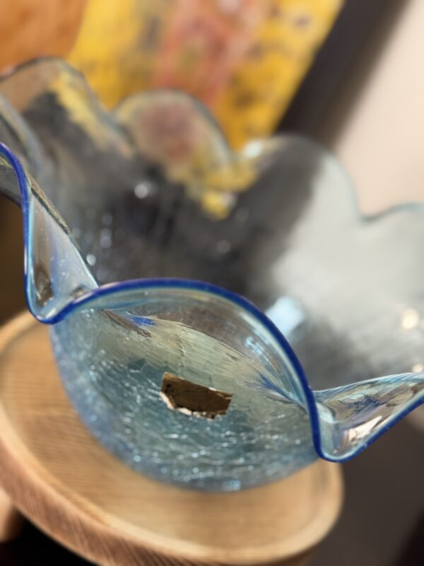 A close-up of a wavy-edged, clear blue glass bowl with a cracked texture, sitting on a wooden surface. The bowl has a blue rim and a small, rectangular label on its side. The background is blurred, featuring yellow and dark tones.