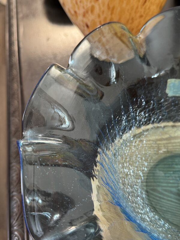 A close-up of a decorative blue glass bowl with scalloped edges and a textured pattern inside. The bowl catches light, creating reflections. In the blurred background, part of a brownish-yellow object and a dark surface are visible.