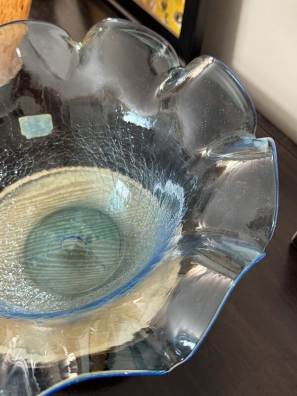 A close-up of a translucent, scalloped-edge glass bowl with a light blue tint sits on a dark wooden surface. The bowl features a textured, crackled pattern inside and partially reflects light from its surroundings.