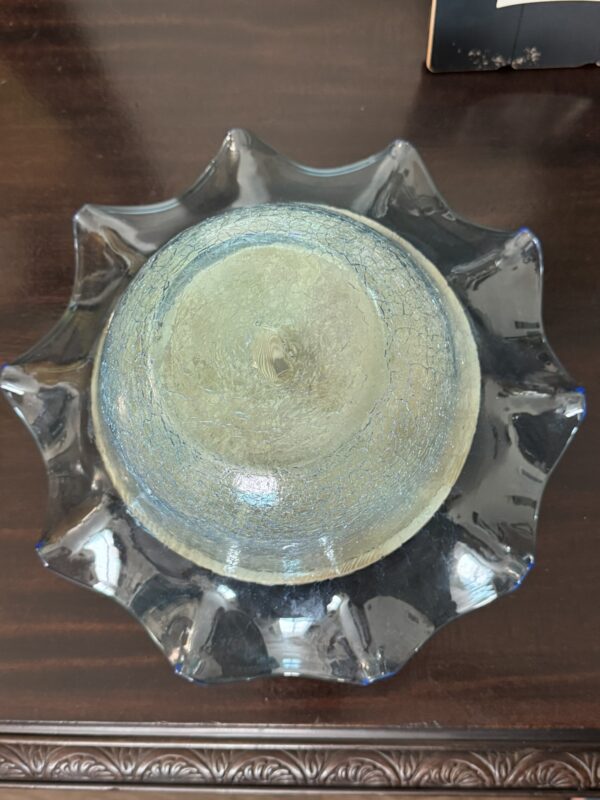 A round glass bowl with a cracked, frosty texture sits upside down on a dark wooden surface. The bowl has a wavy, flared edge and a blue tint, with light reflecting off its smooth, transparent underside. Part of a decorative frame is visible at the top right.