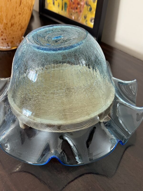 A transparent blue glass bowl sits upside down on a ruffled glass plate, covering a sandwich with white bread on a dark wooden surface. In the background, part of an orange vase and a framed colorful artwork are visible.