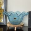 A light blue, wavy-edged glass bowl sits on a round wooden stand with short legs. The bowl has a textured surface and a small gold sticker on the front. It’s placed on a dark tabletop, with framed art partially visible behind it.
