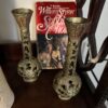 A copy of “Sophie’s Choice” by William Styron stands upright on a wooden shelf, partially framed by two ornate brass vases with patterned openings and colorful crests. The book cover shows a woman’s face and a historical scene.