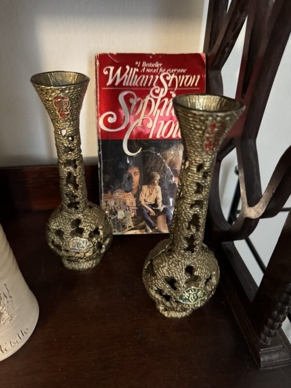 A copy of “Sophie’s Choice” by William Styron stands upright on a wooden shelf, partially framed by two ornate brass vases with patterned openings and colorful crests. The book cover shows a woman’s face and a historical scene.