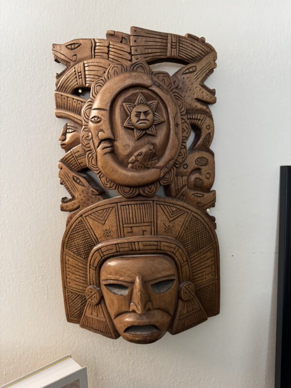 A carved wooden mask features an expressive face with geometric patterns, flanked by a circular motif with a sun symbol and stylized animals above. The intricate design and details reflect Aztec or Mayan artistic influences, set against a light wall.