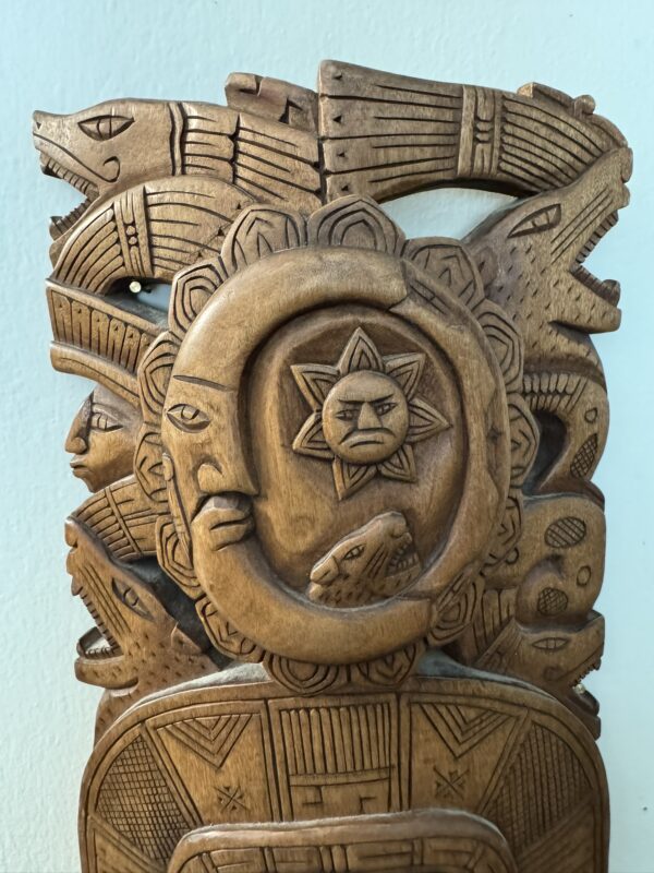 A carved wooden sculpture features Aztec-style faces, animals, and intricate patterns. Central is a circular motif with a stern face inside a sun, surrounded by stylized heads, including a jaguar, an eagle, and geometric designs, all on a light blue background.