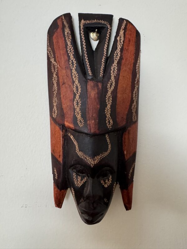 A carved wooden mask with elaborate patterns, featuring dark brown and reddish-orange sections. The mask has a tall, ornate headdress and dotted decorations highlighting the eyes, forehead, and chin. It hangs on a plain white wall.