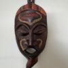 A carved wooden mask with closed eyes, parted lips, and geometric facial patterns in yellow. It features a red and orange striped headdress and a pointed chin detail, hanging on a plain white wall.