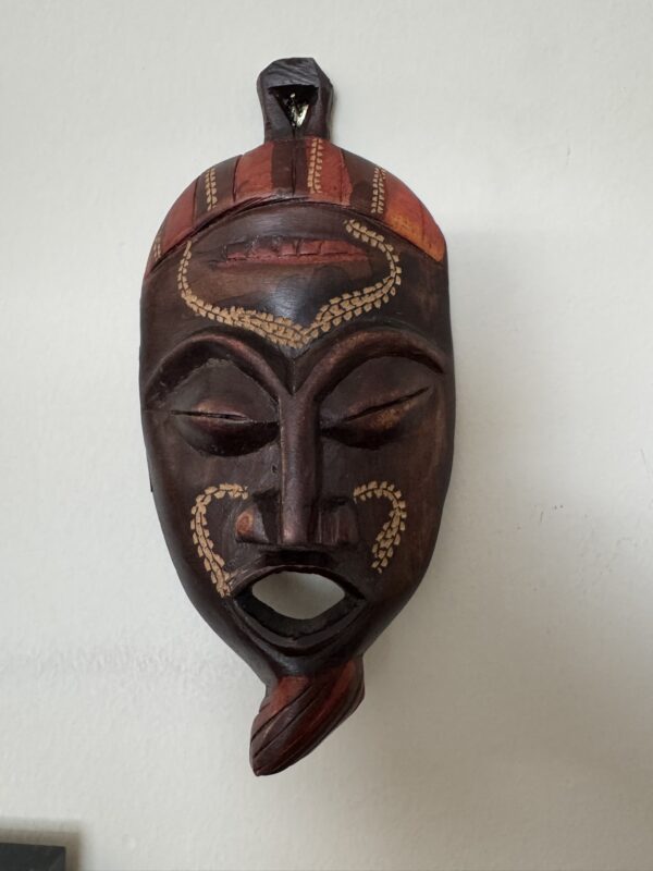 A carved wooden mask with closed eyes, parted lips, and geometric facial patterns in yellow. It features a red and orange striped headdress and a pointed chin detail, hanging on a plain white wall.