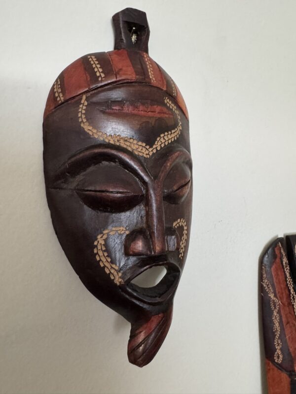 A wooden African tribal mask with a reddish-brown finish, decorated with painted beige dotted patterns and red accents. The mask features closed eyes, high cheekbones, and a long, pointed chin. It hangs on a light-colored wall.