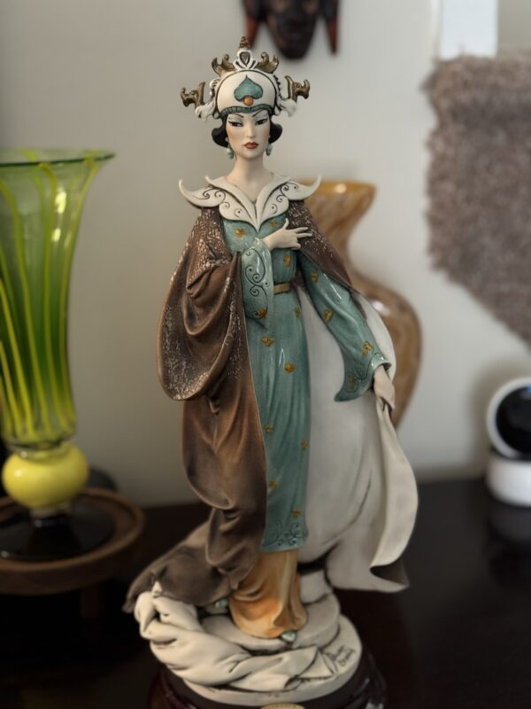 A porcelain figurine of a regal woman in an ornate teal and brown gown with gold accents. She wears a white headdress adorned with antlers and a central blue gem. The figurine stands on a round base, surrounded by glass vases and a security camera.
