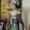 A porcelain figurine of an elegant woman in a teal and brown traditional gown with gold accents, ornate white collar, and elaborate crown. She has black hair, red lips, and holds one hand gracefully to her chest. The background is softly blurred.