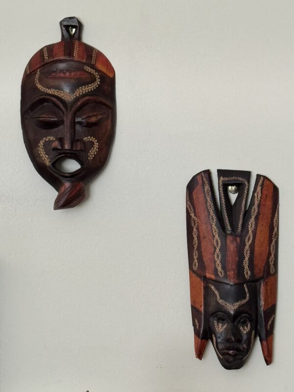 Two decorative wooden masks with intricate carvings hang on a light-colored wall. The left mask has an open mouth and wavy lines, while the right mask features a tall headpiece, both adorned with red and gold-toned painted details.