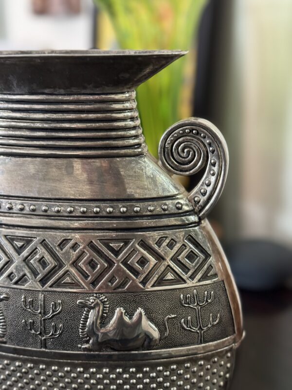 A close-up of an ornate metal vase with engraved patterns, including geometric shapes, a camel, and trees. The vase has a decorative, spiral-shaped handle and a wide, flared rim. The background is blurred with hints of green and black.