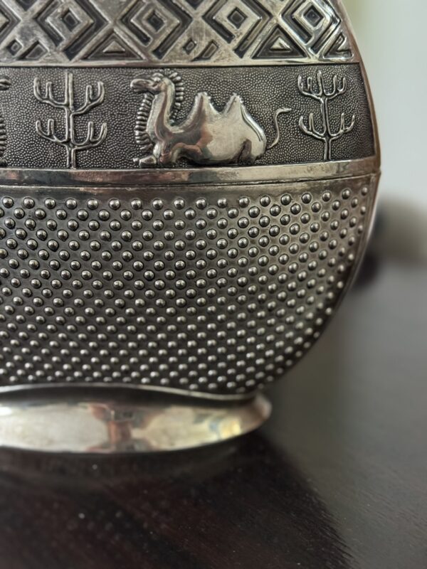 A close-up of a metal decorative object featuring a raised image of a camel and desert plants above a textured pattern of small raised dots, with geometric designs at the top. The object rests on a dark surface.