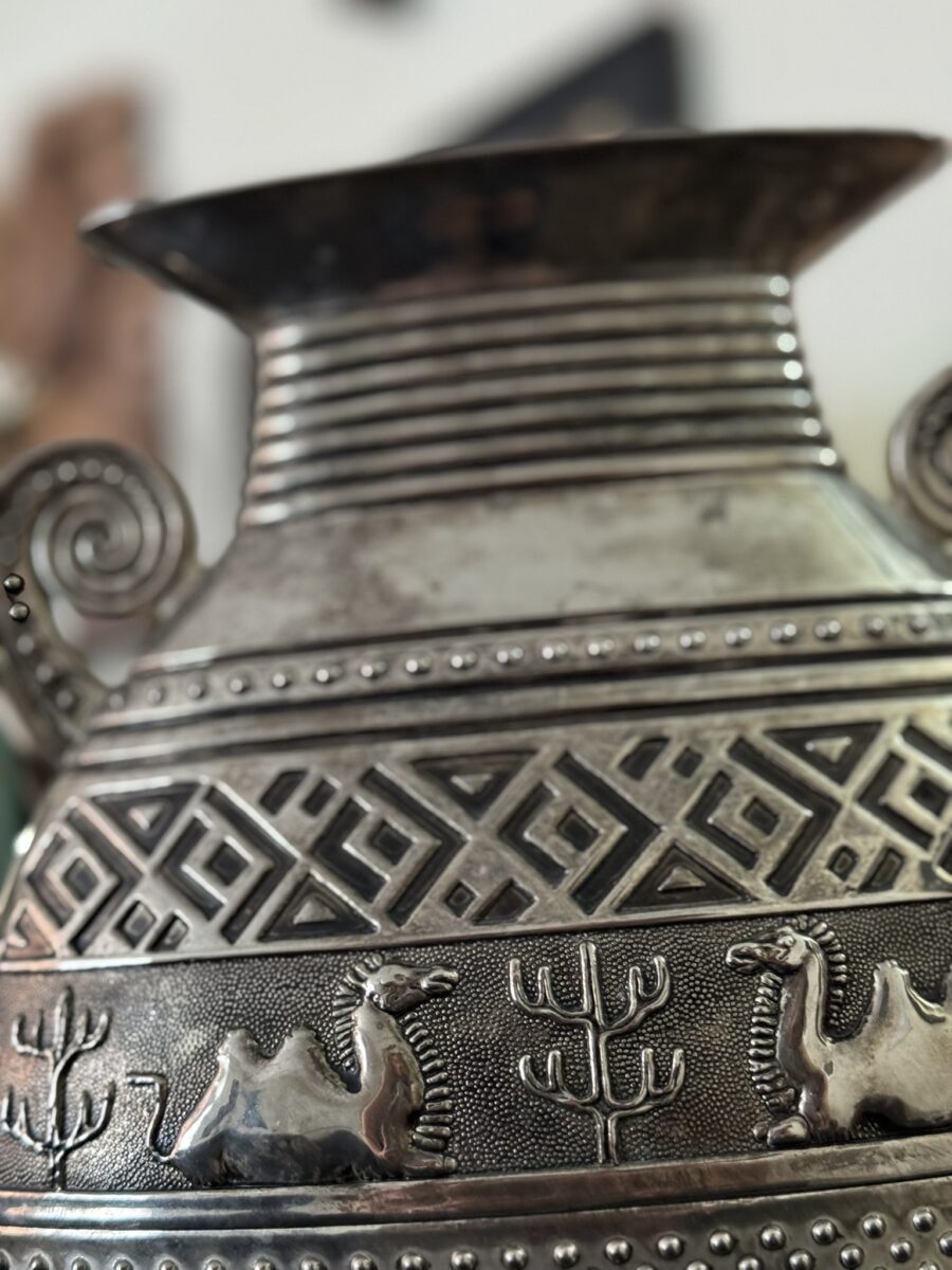 A close-up of a decorative metal vase with intricate engravings. The design features geometric patterns and embossed images of camels and cacti, with spiral-shaped handles visible on the sides. The focus is on the detailed carvings.