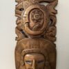 A carved wooden mask with intricate Aztec-style patterns. The top features a sun with a face, surrounded by symbolic animals and geometric designs. The bottom half displays a stern human face with angular features, wearing a detailed headdress.