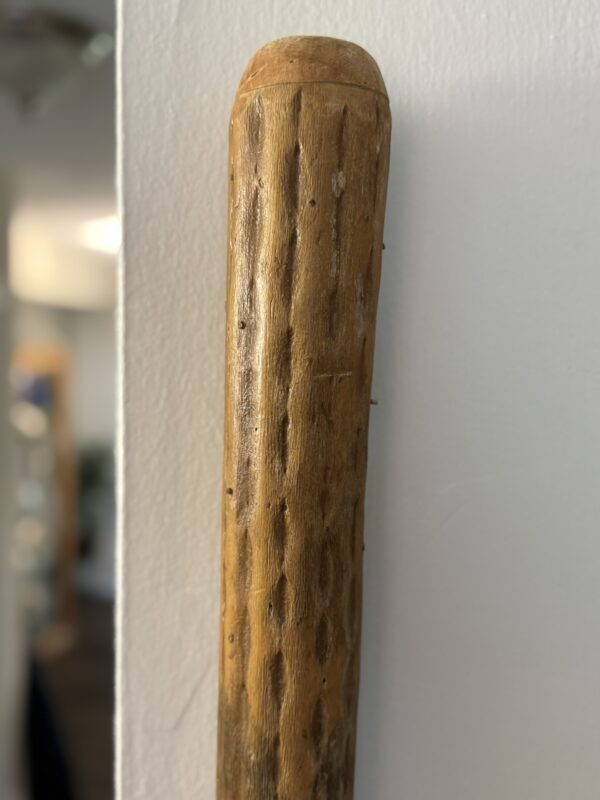 A close-up of a carved wooden stick leaning against a white wall. The wood is smooth with visible grooves, dimples, and a rounded top. The background is softly blurred, showing hints of an indoor setting with light and shadows.
