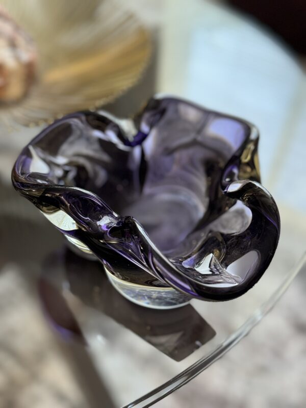 A close-up of a wavy, irregularly-shaped glass bowl with deep purple and black hues, sitting on a glass table. Light reflects off its glossy surface, highlighting the bowls multiple folds and curves. A blurred gold dish is in the background.
