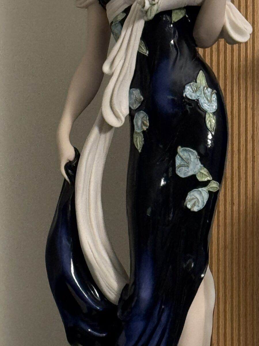 A ceramic figurine of a woman in a dark blue, glossy dress adorned with light blue flowers and green leaves. She holds part of her dress and a cream-colored shawl, against a background of a beige wall and a wood panel. Only her torso and lower body are visible.