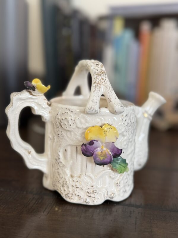 A white ceramic teapot with ornate, textured patterns and a decorative yellow and purple pansy flower on its side sits on a dark wooden surface. In the background, colorful blurred books are visible on a shelf.
