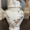 A white porcelain jar with a lid, decorated with a painted bird perched on a flowering branch with pink blossoms and green leaves. The jar has gold accents along the rim and lid, and sits on a wooden surface with blurred background objects.
