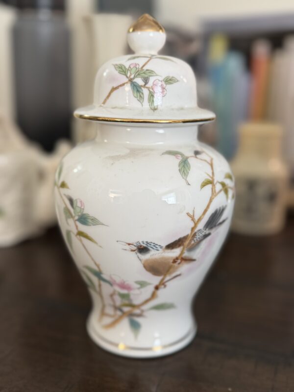 A white porcelain jar with a lid, decorated with a painted bird perched on a flowering branch with pink blossoms and green leaves. The jar has gold accents along the rim and lid, and sits on a wooden surface with blurred background objects.