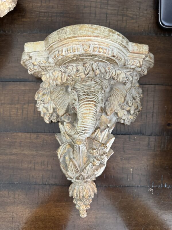 Ornate wall sconce with an elephant head design, featuring detailed carvings of leaves and flowers in a gold-toned finish, resting on a dark wooden surface.
