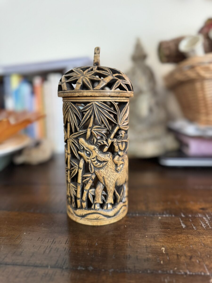 A carved cylindrical container with a lid, featuring intricate designs of a deer among bamboo plants. The wooden piece sits on a dark wooden table, with blurred household items and books in the background.