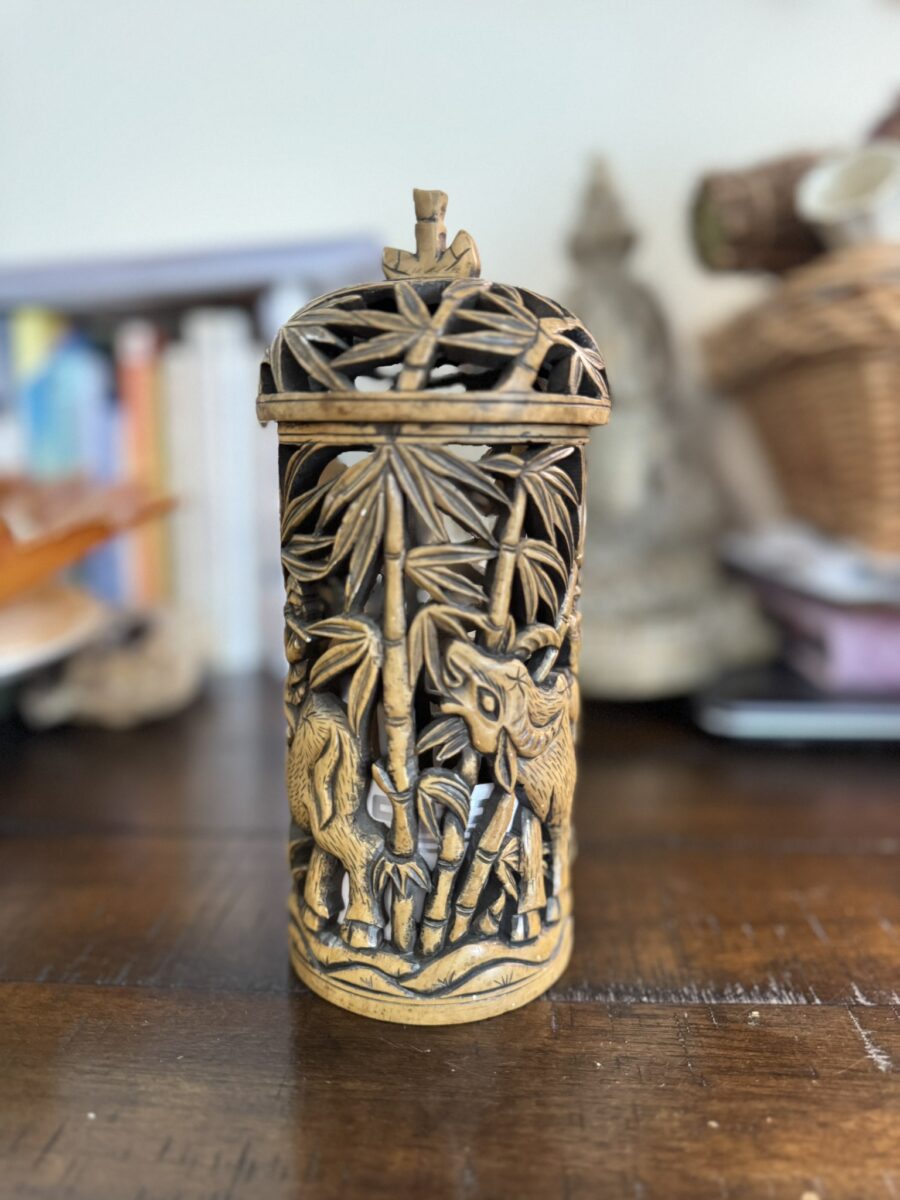 A carved wooden cylindrical container with a lid, featuring detailed deer and bamboo designs. The background shows blurred books, a wicker basket, and decorative objects atop a polished wooden surface.