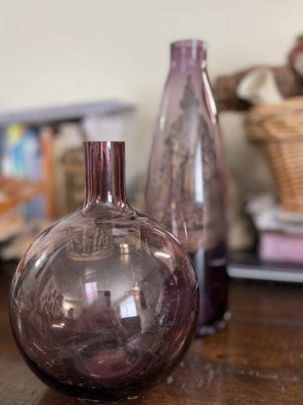 Two purple glass vases sit on a wooden table. One vase is round and squat with a narrow neck, while the other is taller and slimmer with an etched design. In the background, there are blurred books, a woven basket, and other household items.