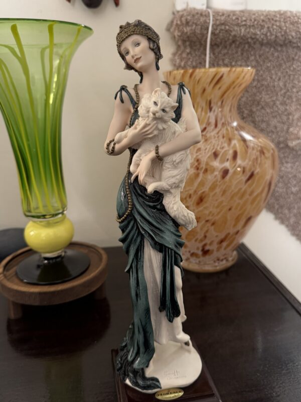 A porcelain figurine of a woman in a green gown and headpiece holds a fluffy white cat. She has a calm expression. Behind her are a tall green glass vase with a yellow base and a marbled orange vase, all on a dark wooden surface.