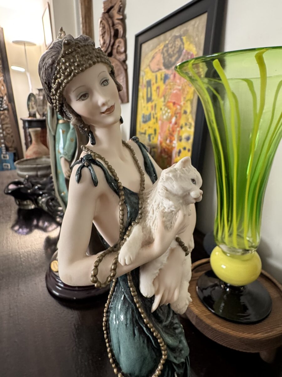 A detailed porcelain figurine of a woman in a green dress and beaded headdress holds a white cat. The figurine is on a dark wooden surface next to a tall green glass vase and a framed artwork in the background. The setting appears decorative and elegant.