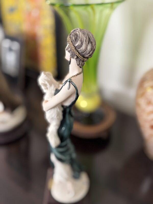 A porcelain figurine of a woman in a dark green dress holding a white cat. The woman has short, wavy brown hair with a gold headband. She stands on a dark surface with a large green vase and other decor blurred in the background.