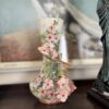 A decorative ceramic vase sits on a dark surface. The vase is adorned with textured pink flowers, green leaves, and brown branches. Behind it is a blurred framed artwork and part of a statue in a green dress is visible on the right.