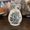 A cream-colored ceramic vase with a narrow neck, decorated with detailed black floral patterns, sits on a dark wooden surface. The background is softly blurred, showing books and various decorative objects.