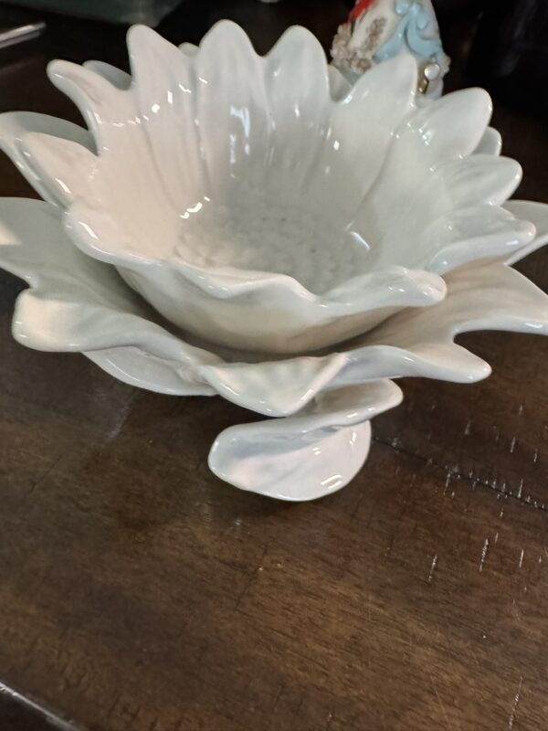 A white ceramic bowl shaped like a flower with layered petal edges sits on a dark wooden surface. The flower-shaped bowl is elevated on a matching pedestal base, echoing the petal design. Part of a decorative object is visible in the blurred background.