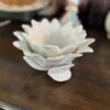 A white ceramic bowl shaped like a flower with layered petals sits on a wooden surface. The bowl has a small leaf detail at the base. In the blurred background are additional decorative items, including a figurine and another dish.