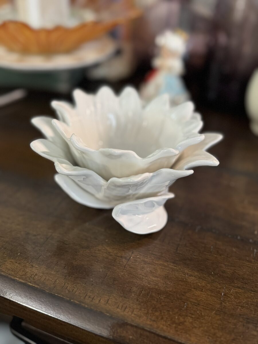 A white ceramic bowl shaped like a flower with layered petals sits on a wooden surface. The bowl has a small leaf detail at the base. In the blurred background are additional decorative items, including a figurine and another dish.