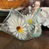 A small, wavy-edged glass dish with painted white daisies featuring yellow and orange centers sits on a wooden surface. In the background, there are books, a decorative amber glass plate, a small figurine, and a porcelain item.