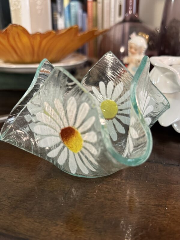 A small, wavy-edged glass dish with painted white daisies featuring yellow and orange centers sits on a wooden surface. In the background, there are books, a decorative amber glass plate, a small figurine, and a porcelain item.