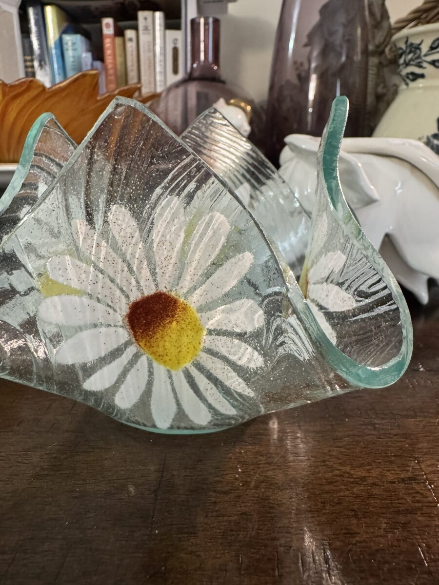 A decorative glass bowl with wavy edges features white daisies with yellow and orange centers painted on its sides. The bowl sits on a wooden table, surrounded by various ceramic and glass objects, with books visible in the background.