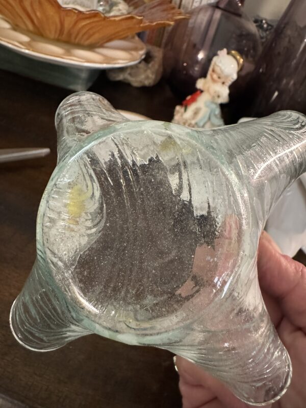 A hand holds up a clear glass object with four rounded, curved legs at the base. The glass is slightly wavy and has visible bubbles and imperfections. In the background, there are decorative items, including a porcelain figurine and a brown vase.