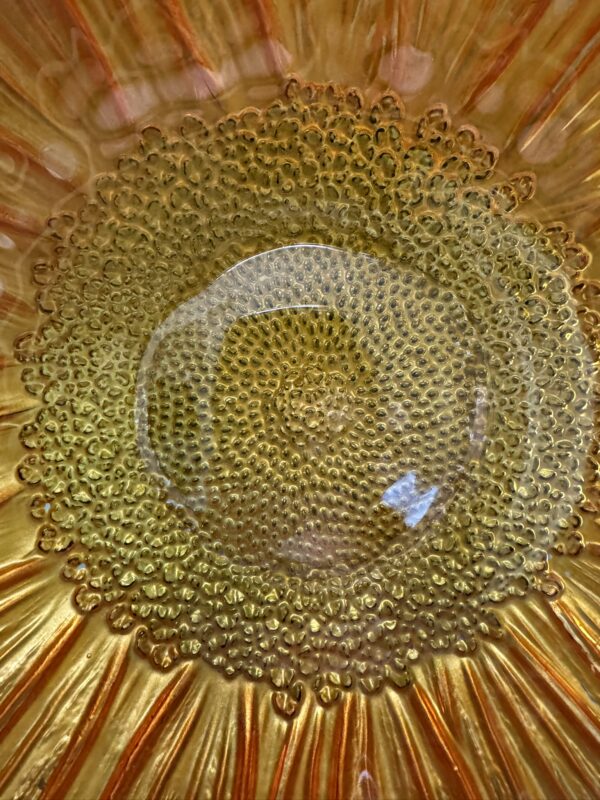 A close-up of a decorative glass plate with an intricate, raised dotted pattern in the center. The plate radiates orange and gold tones outward, resembling a sunflower or sunburst. Light glints off the glass, emphasizing its texture and translucent quality.