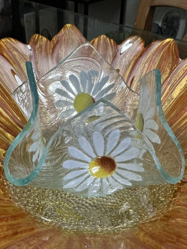 A decorative glass bowl with scalloped edges, orange and yellow hues, and a textured base holds a smaller, clear glass vase with blue-tinted edges and white daisy flower designs with yellow centers. The setup is on a reflective glass surface.