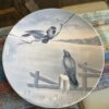 A decorative plate with a monochrome blue-gray illustration shows two birds, one perched on a branch and the other on a wooden fence post, against a rural landscape with a lake, grassy field, and partly cloudy sky in the background.