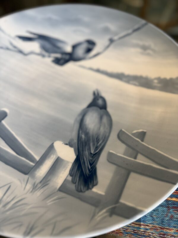A decorative plate with a blue-gray illustration of two birds: one perched on a wooden fence in the foreground, and another flying toward a branch above, set against a serene landscape background. The plate rests on a colorful woven surface.