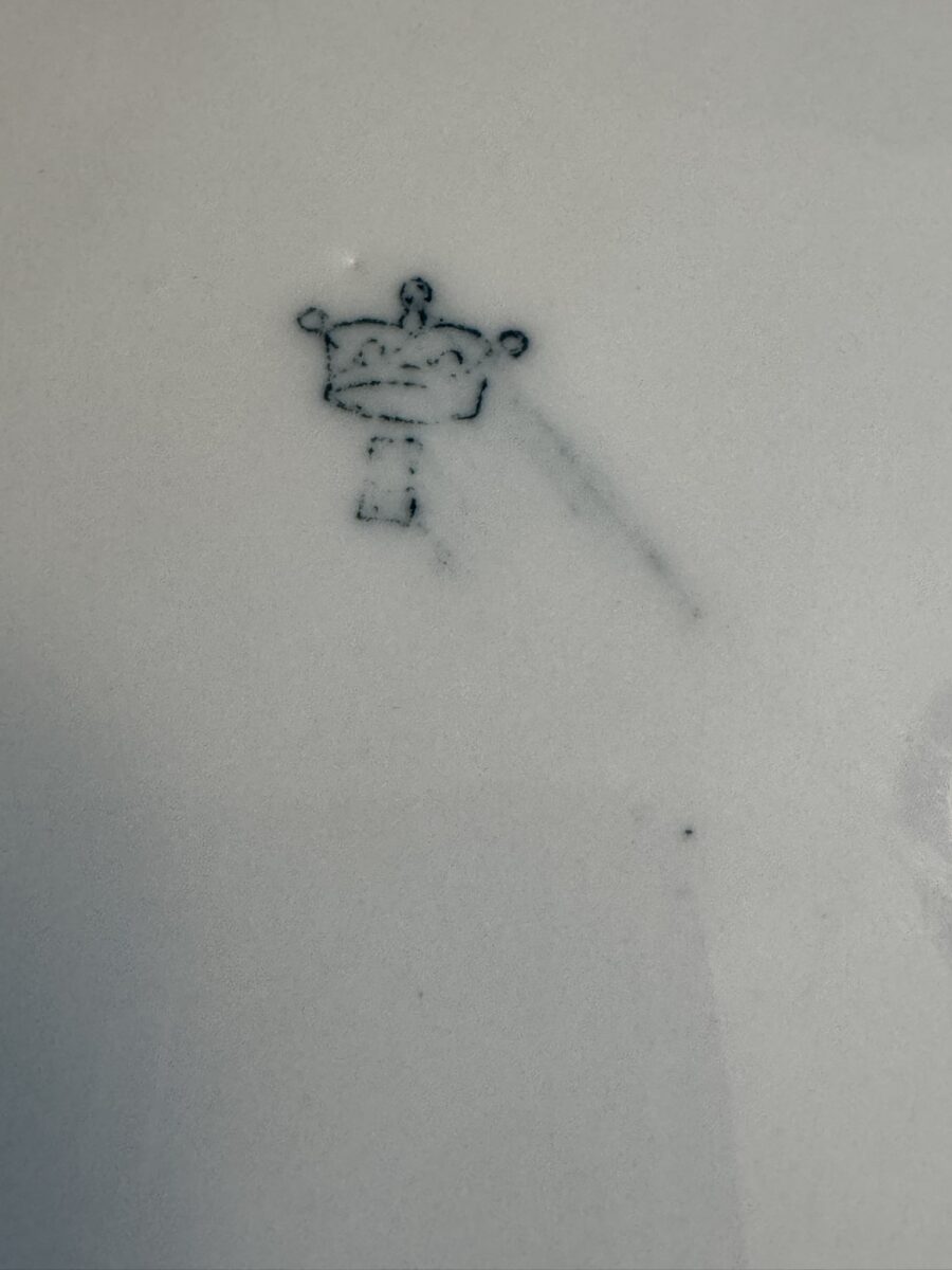 A close-up of a faint blue mark on white porcelain. The mark resembles a stylized crown with three circular points on top and a small rectangular shape below it. The lines appear dotted and slightly smudged, and the surface is smooth with some light marks.