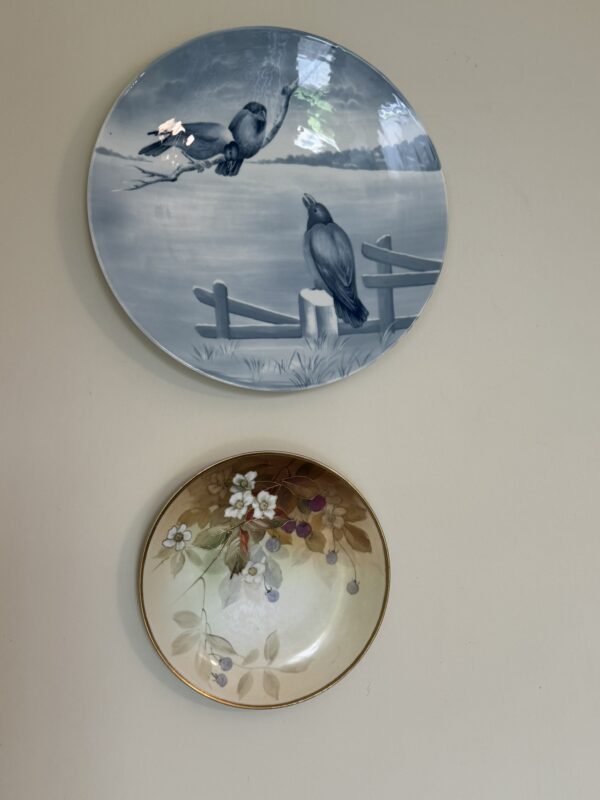 Two decorative plates hang on a pale wall. The top plate features two black birds perched on a wooden fence by a lake, drawn in blue-gray tones. The lower plate shows painted leaves, flowers, and berries in soft, earthy colors.
