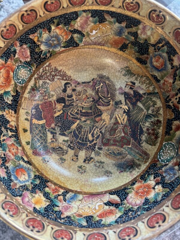 A decorative ceramic bowl with a crackle glaze shows a colorful scene of samurai and figures in traditional Japanese attire, surrounded by floral patterns in red, blue, orange, and gold on a black background along the rim.